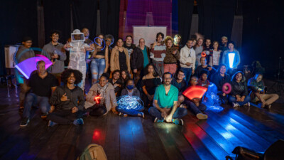 Alunos da Oficina de Light-Painting e Animação realizado no Tendal da Lapa conversam e trocam experiências enquanto manuseiam câmeras em um ambiente com pouca iluminação.