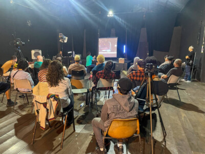 Alunos da Oficina de Light-Painting e Animação realizado no Tendal da Lapa conversam e trocam experiências enquanto manuseiam câmeras em um ambiente com pouca iluminação.