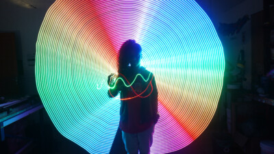 Uma figura sombreada está diante de um círculo vibrante de luzes em espiral nas cores do arco-íris, com traços luminosos adicionais ao redor da tela.