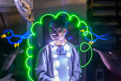 Um jovem com cabelo escuro está no centro, cercado por luzes neon verdes onduladas e figuras de pássaros azuis. Mãos desfocadas aparecem nas laterais.